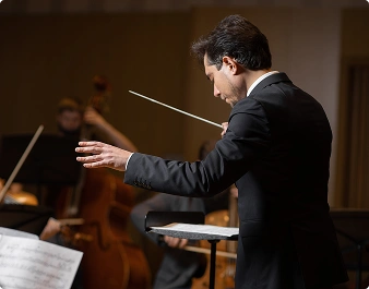 Dyrygent w ciemnym garniturze z pasją prowadzi orkiestrę. W prawej ręce trzyma batutę, gestykulując ekspresyjnie. W tle muzycy grają na instrumentach smyczkowych.