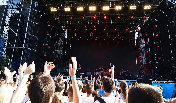 Scena koncertowa z dużą publicznością zwróconą w stronę jasno oświetlonej sceny. Czerwone światła oświetlają scenę, tworząc energiczną atmosferę. Widzowie podnoszą ręce.