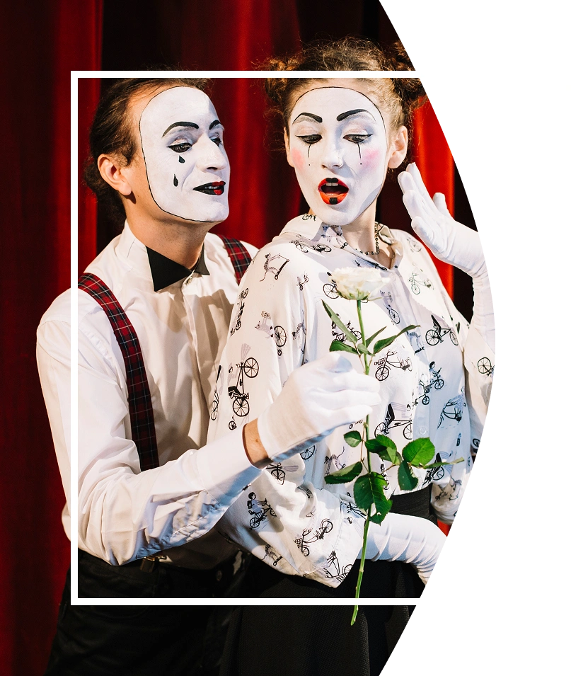 Dwóch mimów z pomalowanymi na biało twarzami i wyrazistym makijażem występuje przed czerwonymi zasłonami. Jeden z nich podaje białą różę, wywołując zaskoczenie i elegancję.