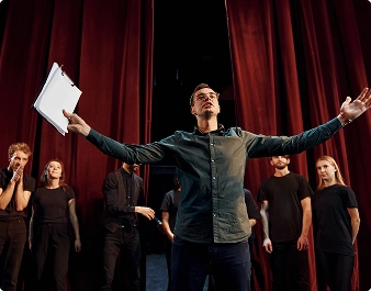 Osoba stoi przed czerwoną kurtyną teatralną z wyciągniętymi rękami, trzymając w nich notes. Kilka innych osób w czarnych ubraniach obserwuje ją, wyglądając na skupionych i podekscytowanych.