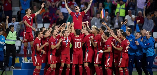 Drużyna piłkarska w czerwonych koszulkach świętuje zwycięstwo, podnosząc gracza trzymającego trofeum na podium. W tle kibice i trenerzy radośnie dopingują.