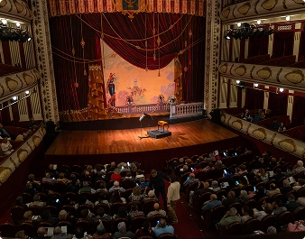 Wnętrze wielkiego teatru z wzorzystymi czerwonymi zasłonami, ozdobnymi kremowymi balkonami i publicznością siedzącą na widowni. Ciepła atmosfera pełna oczekiwania.